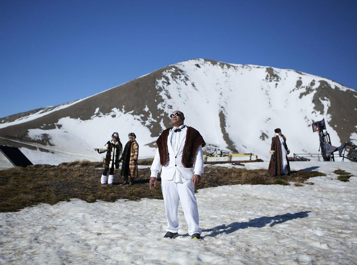 In. this still of Christopher Ulutupu's video work, the artist's father stands on a sunny, snowy mountain peak wearing a dapper white suit, sunglasses and fur throw, looking up towards the shining sun. The artist’s mother and sisters are in the background, all wearing glamorous clothing in the bright snow