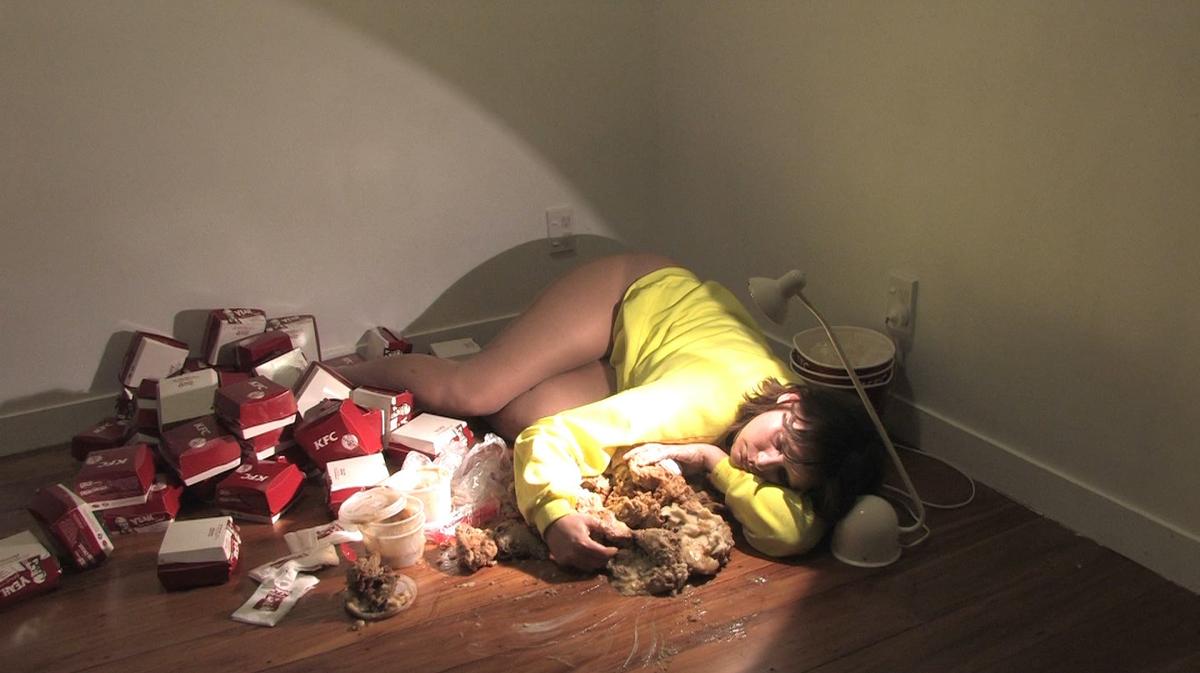 In the corner or an empty room. Sarah Jane lies down sleepily cuddling a large pile of KFC chicken and potato and gravy