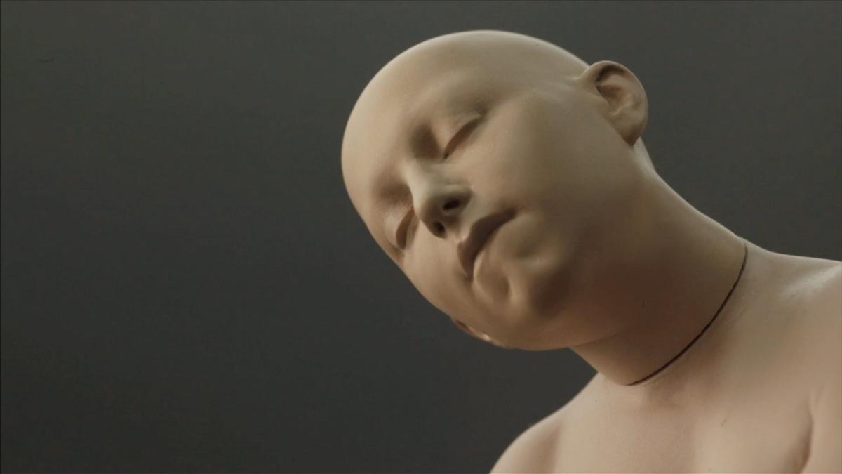 A computer generated person or doll is shown in soft lighting with their eyes closed and head titled upwards looking peaceful