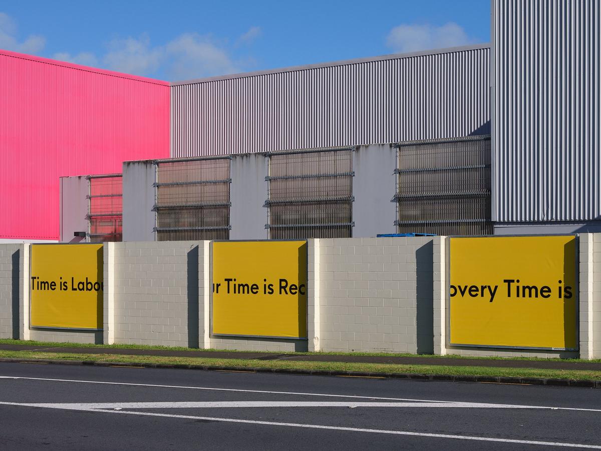 Three billboards with black text on a yellow background read "recovery time is labour time," affixed to the exterior of a steel building with one bright pink wall.