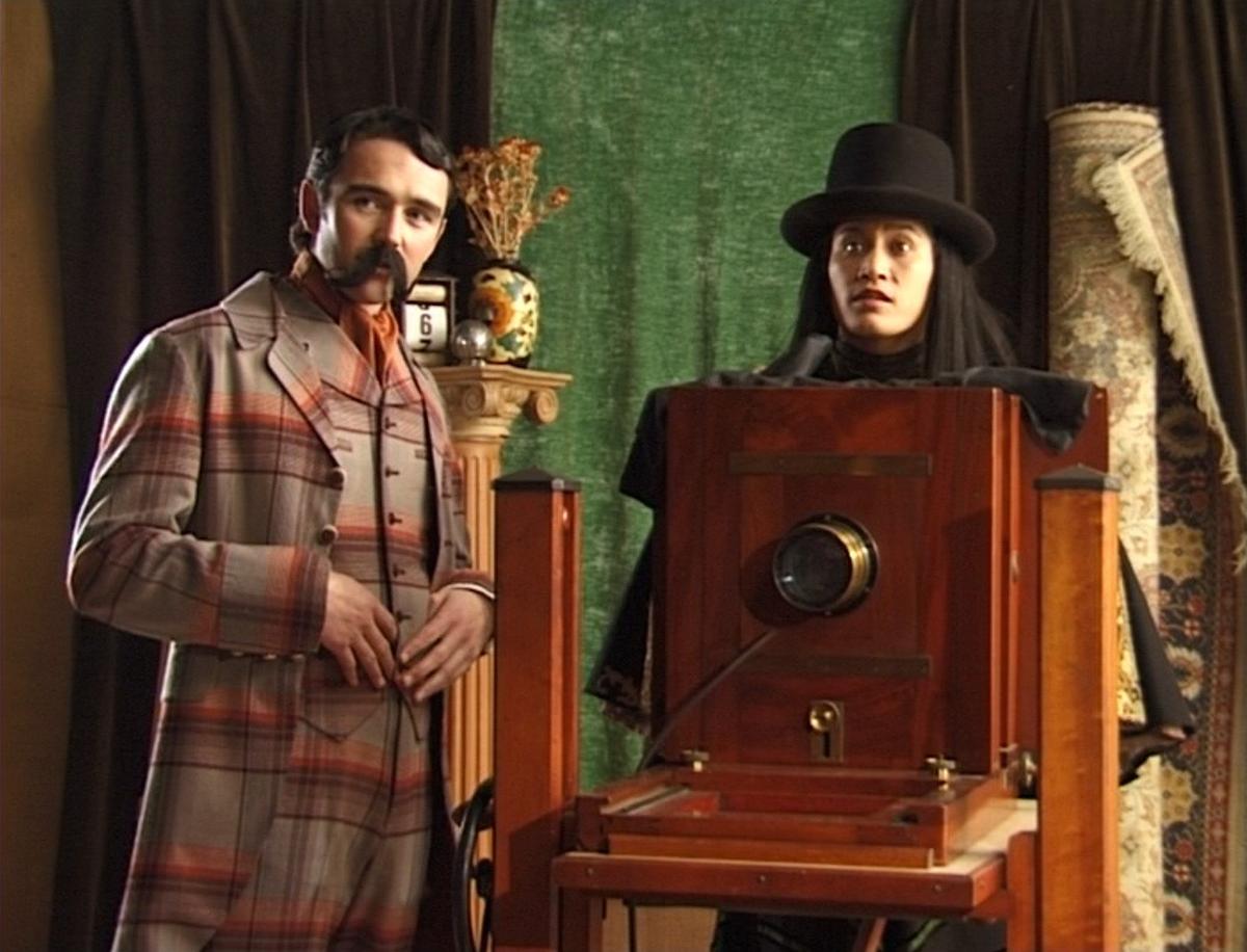 In a colonial photographers studio a comically mannered British gentleman and a Māori photographer stand by a large format camera