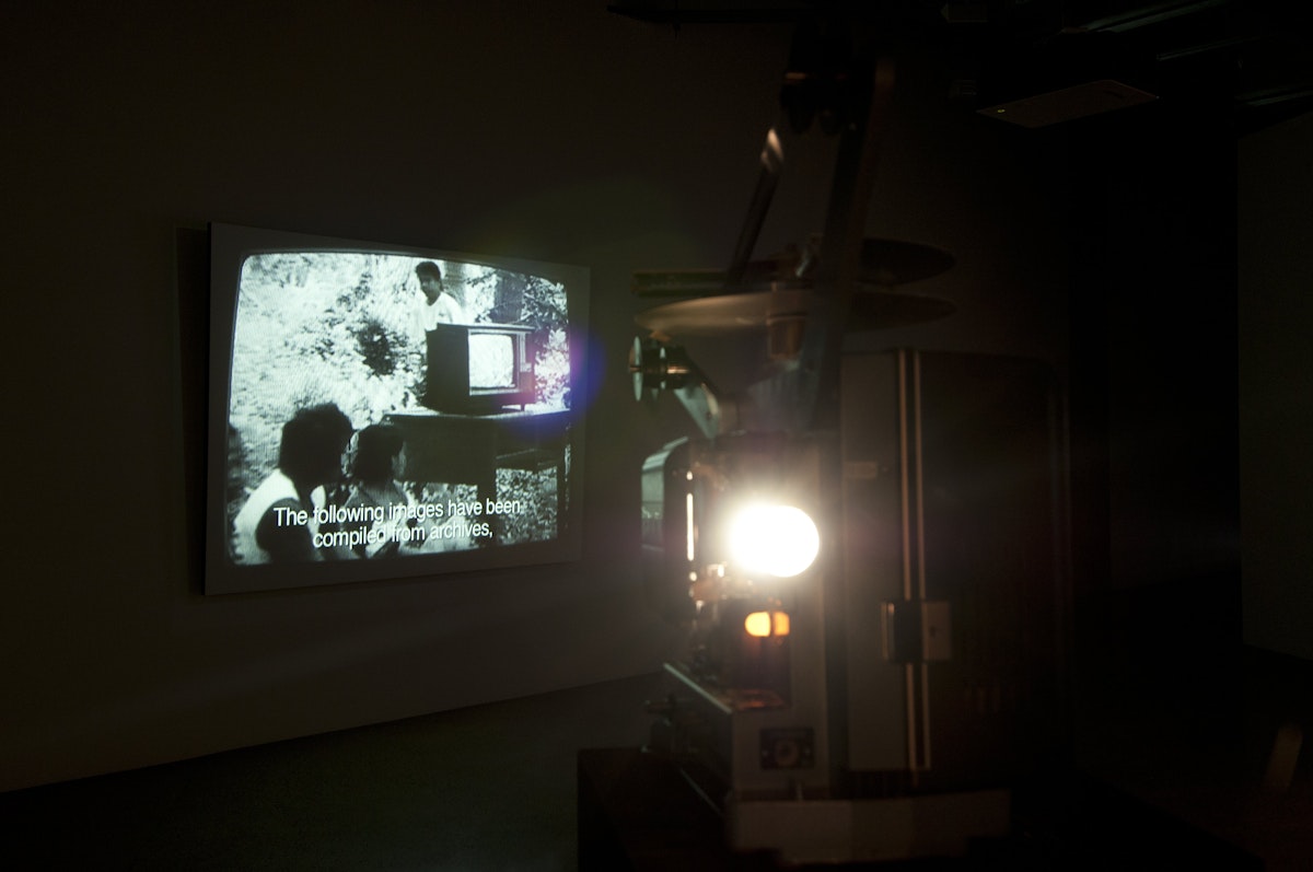 A film projector throws an image onto the wall of people near a tv, the subtitles read "the blowing up of images has been complied from archives"