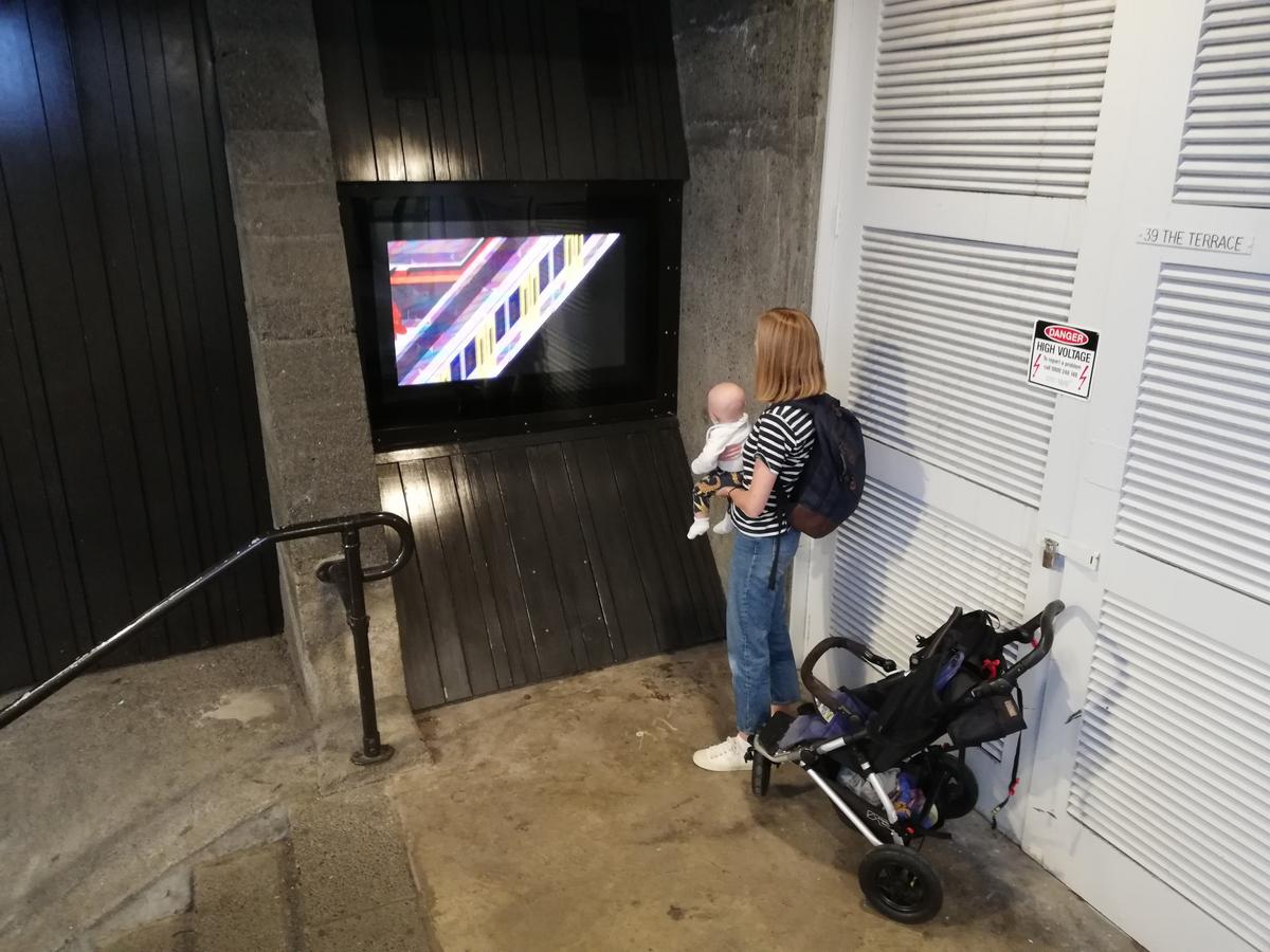 A person stands next to a stroller, holding a small baby and watching the video work playing on masons screen