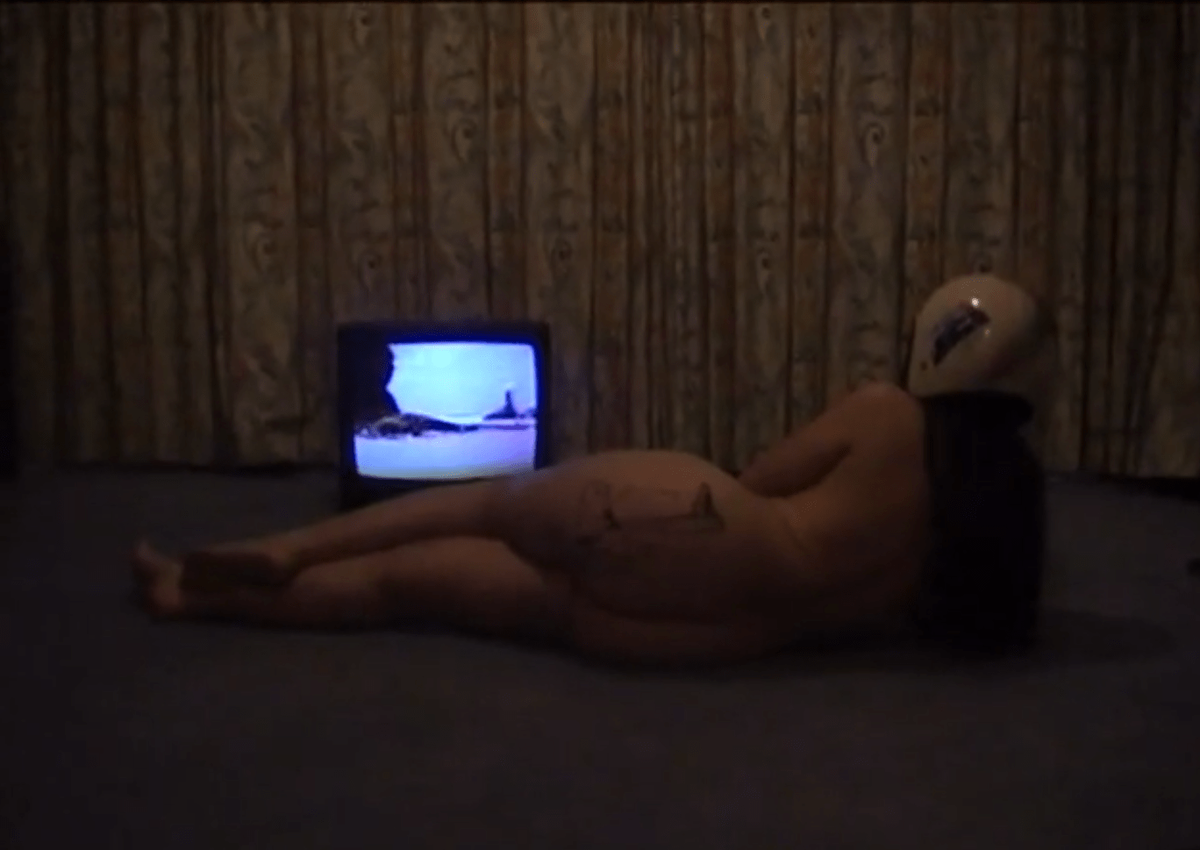 A naked woman wearing a helmet lies on the carpet in an almost empty room watching a small box tv