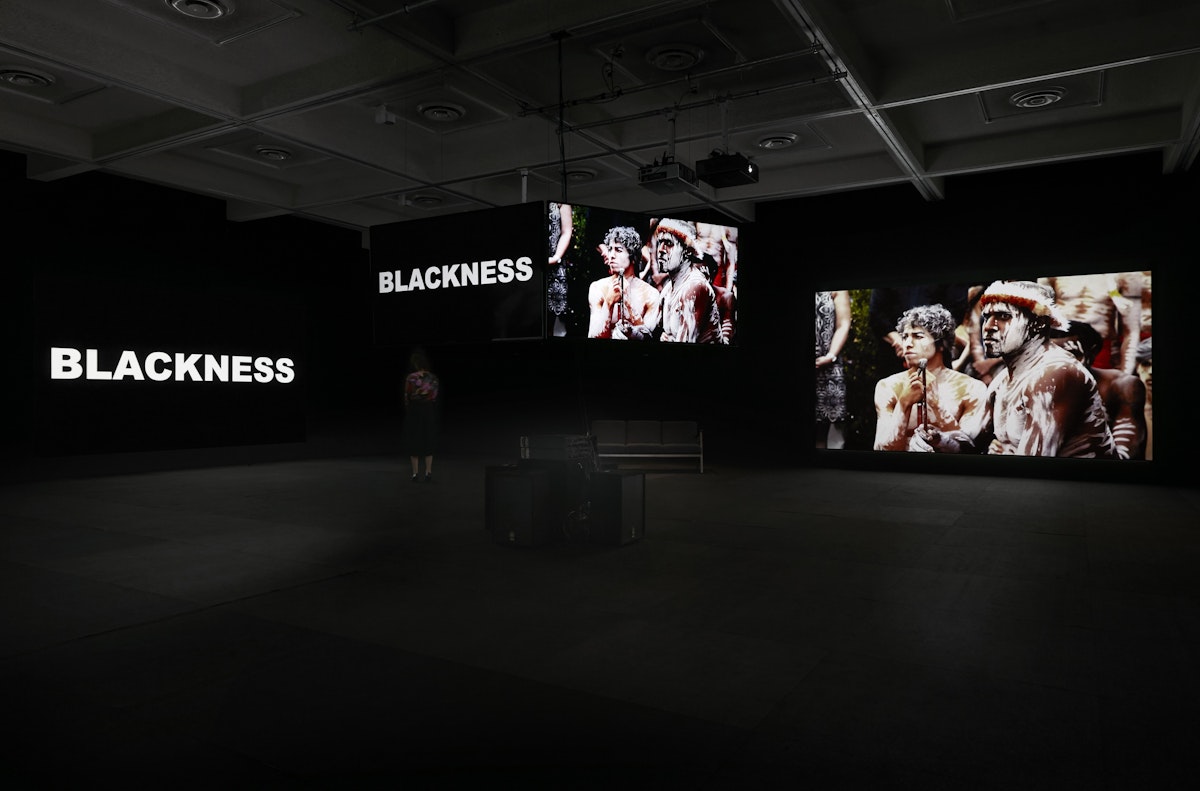 Four large screens are suspended in a dark gallery, held at multiple heights. Two screens have the word "BLACKNESS" displayed and two screens show footage of aboriginal people in traditional dress