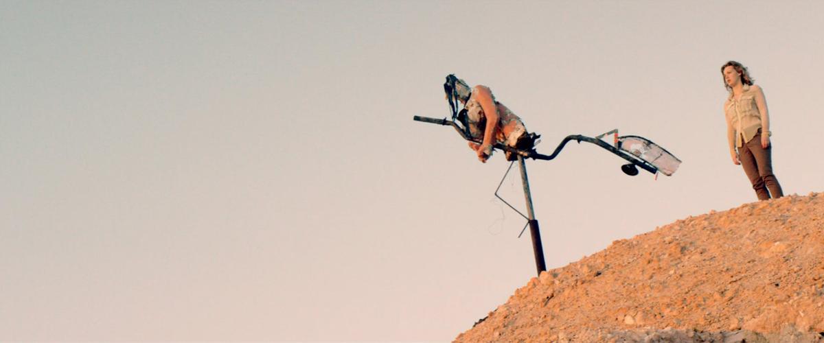 A cyborg and a person stand on a small hill of dry red earth