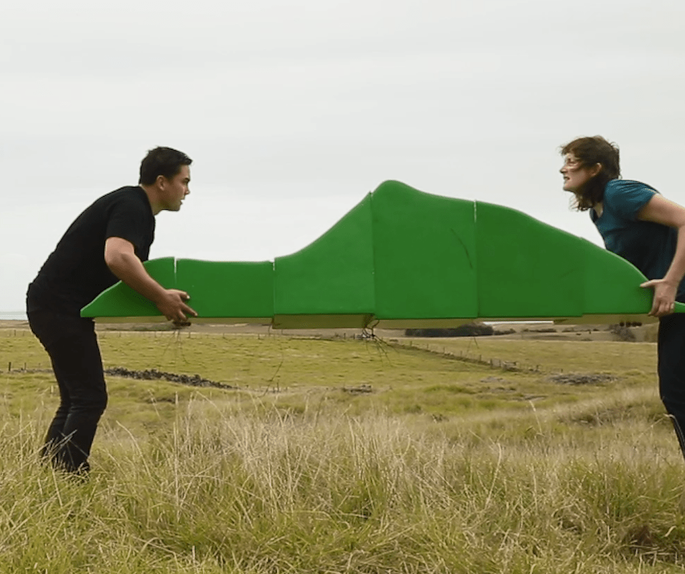 Rebecca Hobbs and Martin Awa Clarke Langdon hold a mountain like green sculpture between them whilst standing in a grassy field