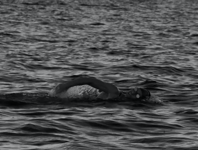 A professional swimmer is moving their body through water, we see them taking a large breath.