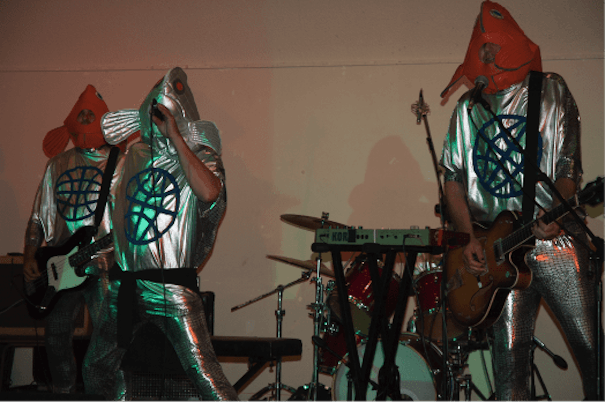 Three people perform music wearing shiny silver costumes and fish masks