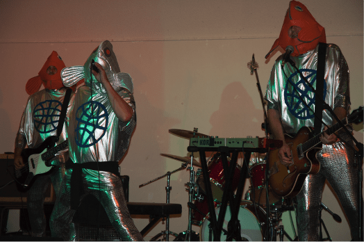 Three people perform music wearing shiny silver costumes and fish masks