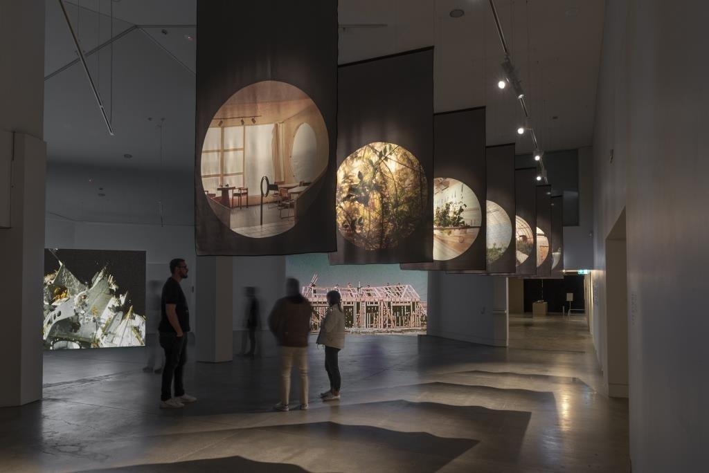 Three people stand in a gallery which displays two large screens playing video works as well as eihgt large scale photographic prints on fabric which are hung above the audiences head from the ceiling.