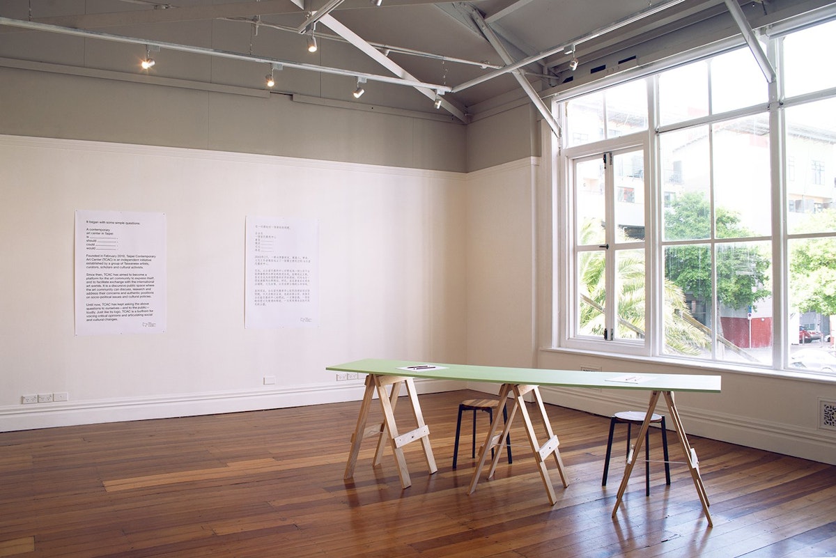 Bright light streams through large windows into a gallery with woooden floors, two large text images are on the wall and there is a table in the foreground