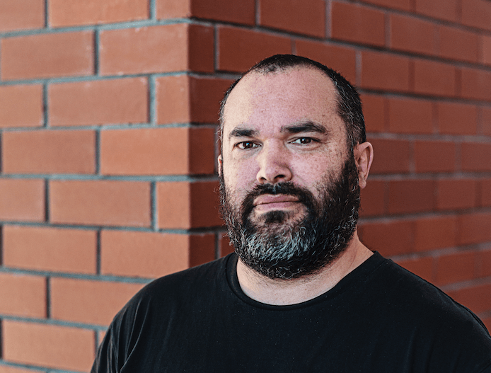 Rangituhia wears a black t-shirt and has a dark beard, he looks at the camera with a serious expression