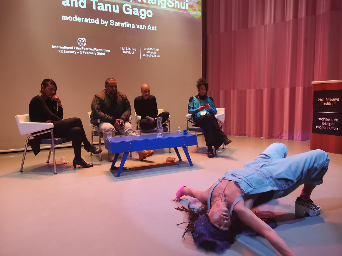 Four people sit a panel in the background whilst a performer in the foreground stretched their body across the floor