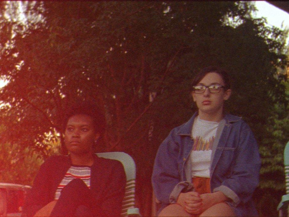 Teenagers sit on deck chairs on the pavement looking out into the distance with bored expressions