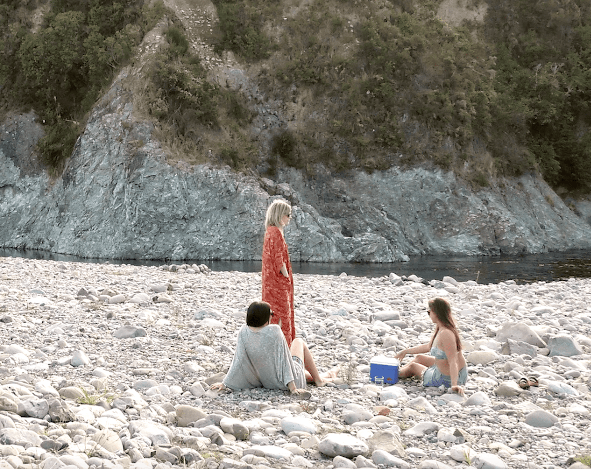 Three pākehā people walk around and picnic on river rocks