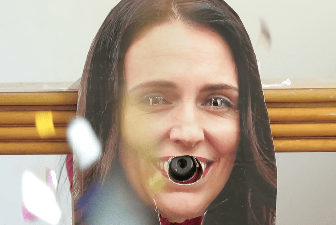 A cardboard cut-out of Jacinda Ardern has had its eyes and mouth cut out. A confetti canister is poking out of her mouth, exploding glitter