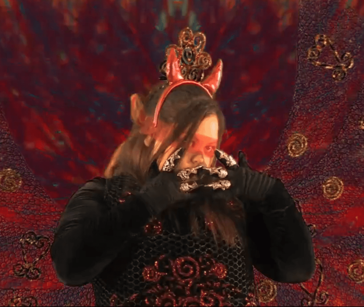 Candice is dressed in glamorous costuming, wearing black gloves with jewellery on top, devil horns and a red and black dress. They are dancing in front of a swirling red textured background