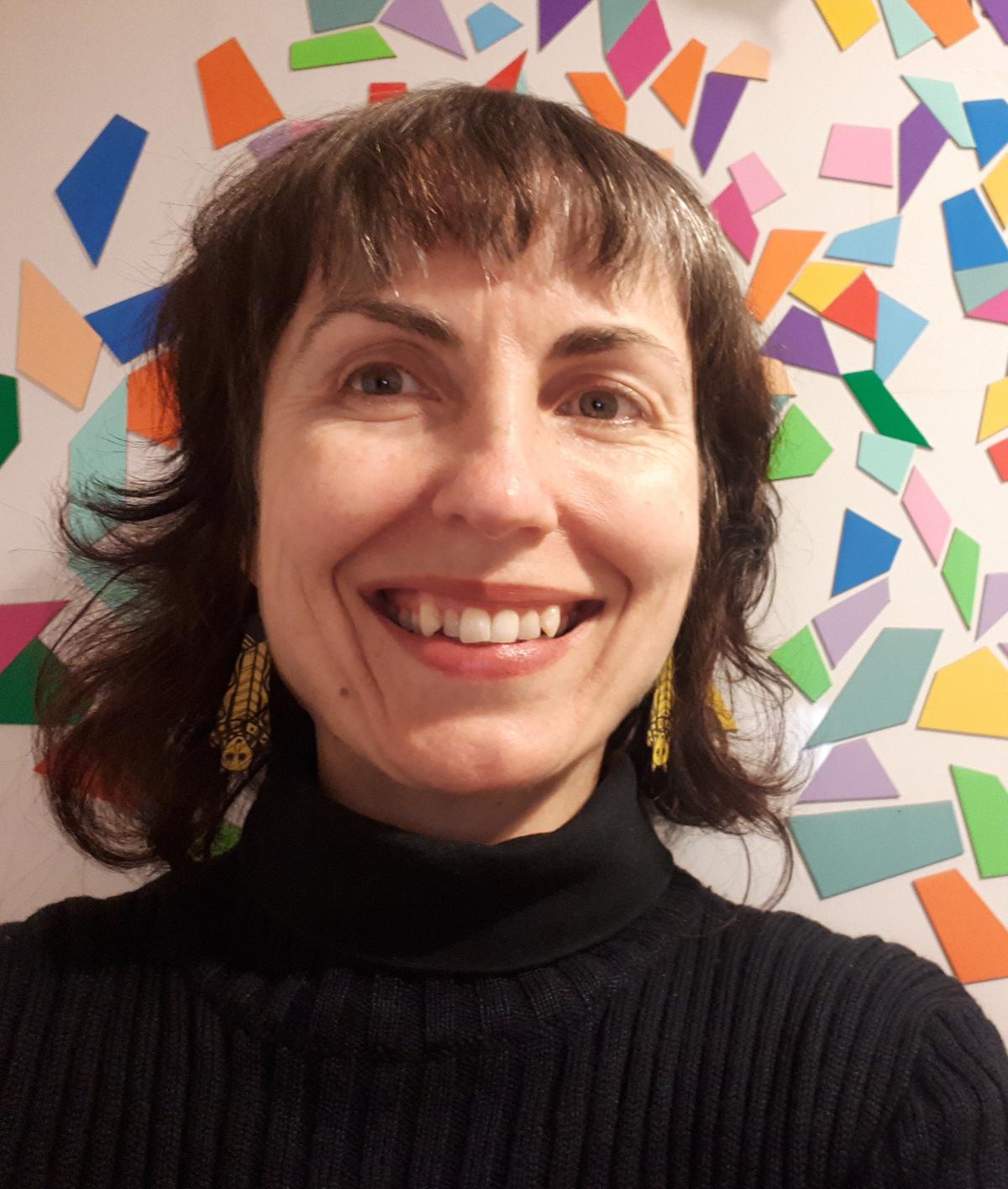 A woman smiles for the camera, behind her are colourful mosaic tiles stuck to the wall.