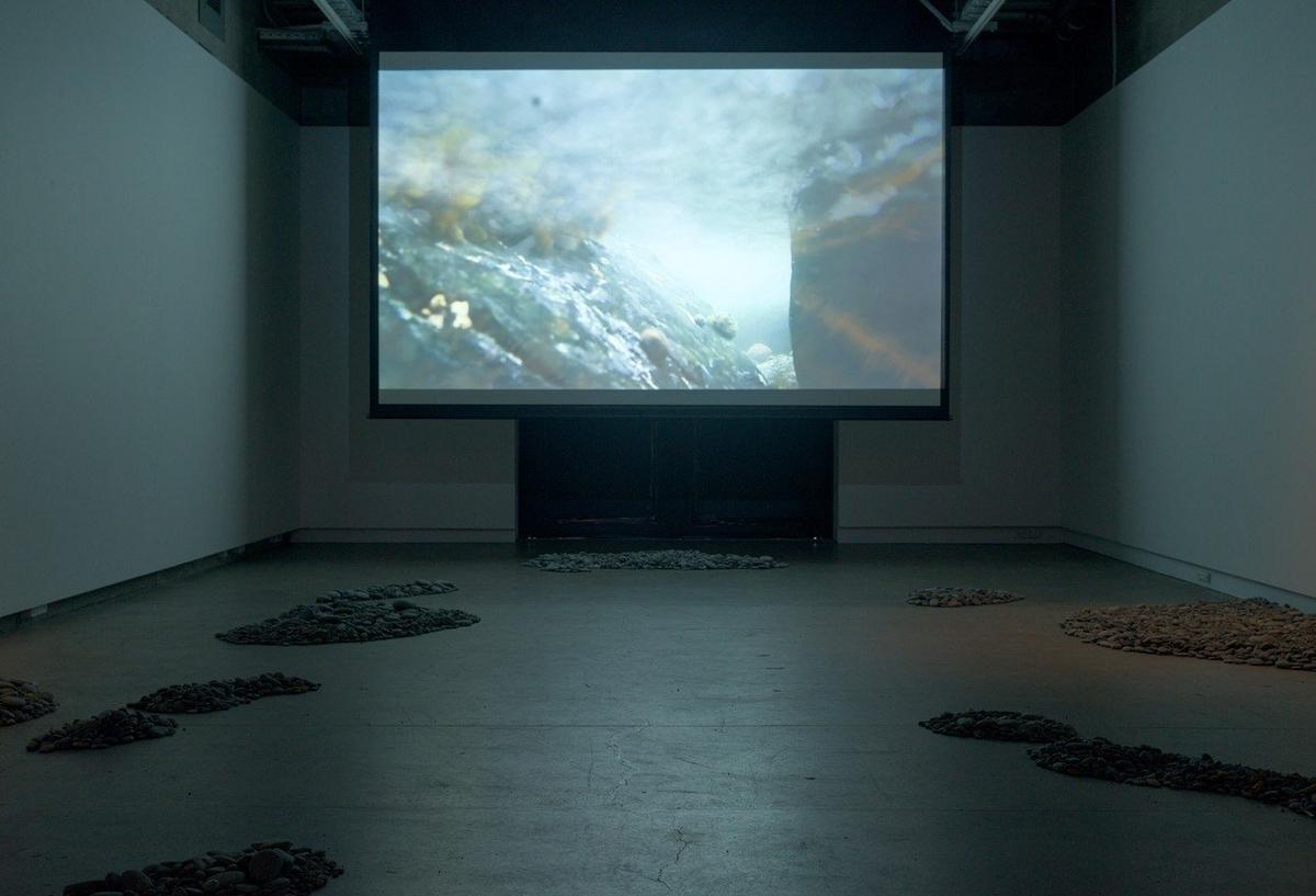 A screen plays an underwater image in a darkened room. There are shaped piles of stones in the foreground.