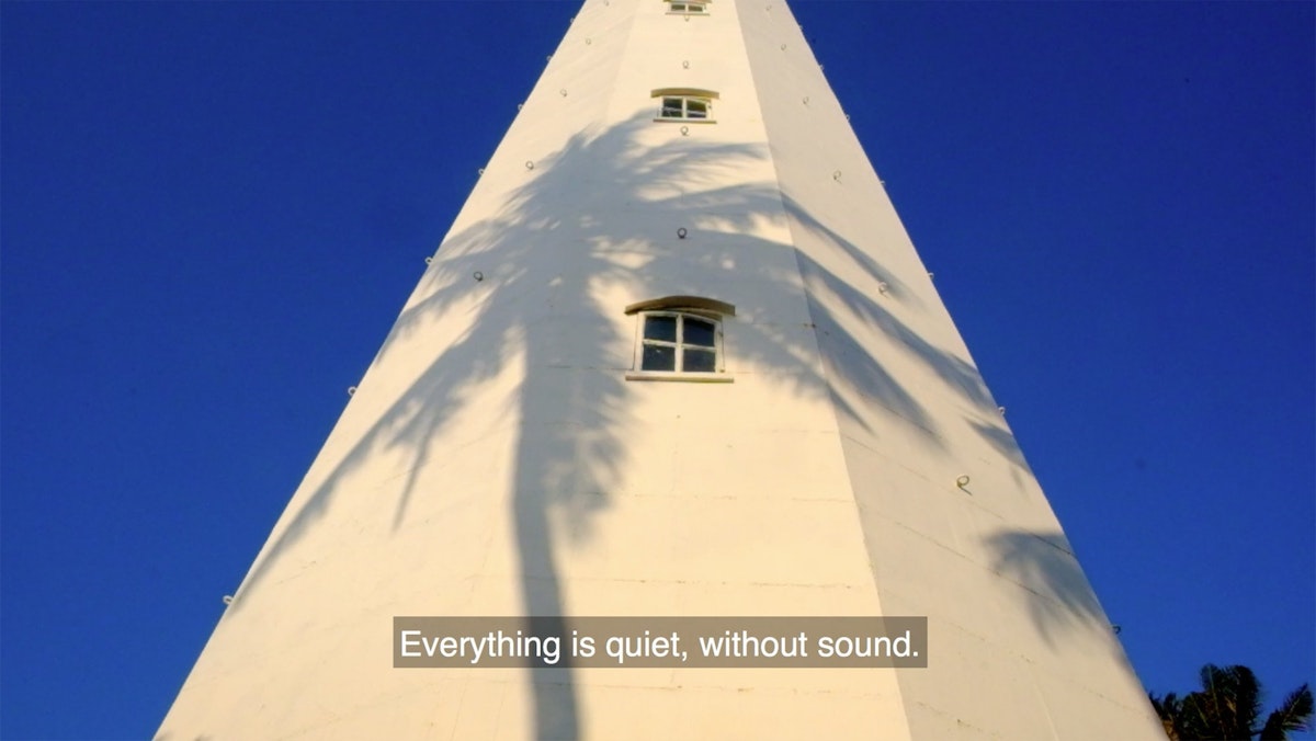 The shadow of a palm tree is exposed on large white tower, subtitles on the image say "Everything is quiet, without sound."