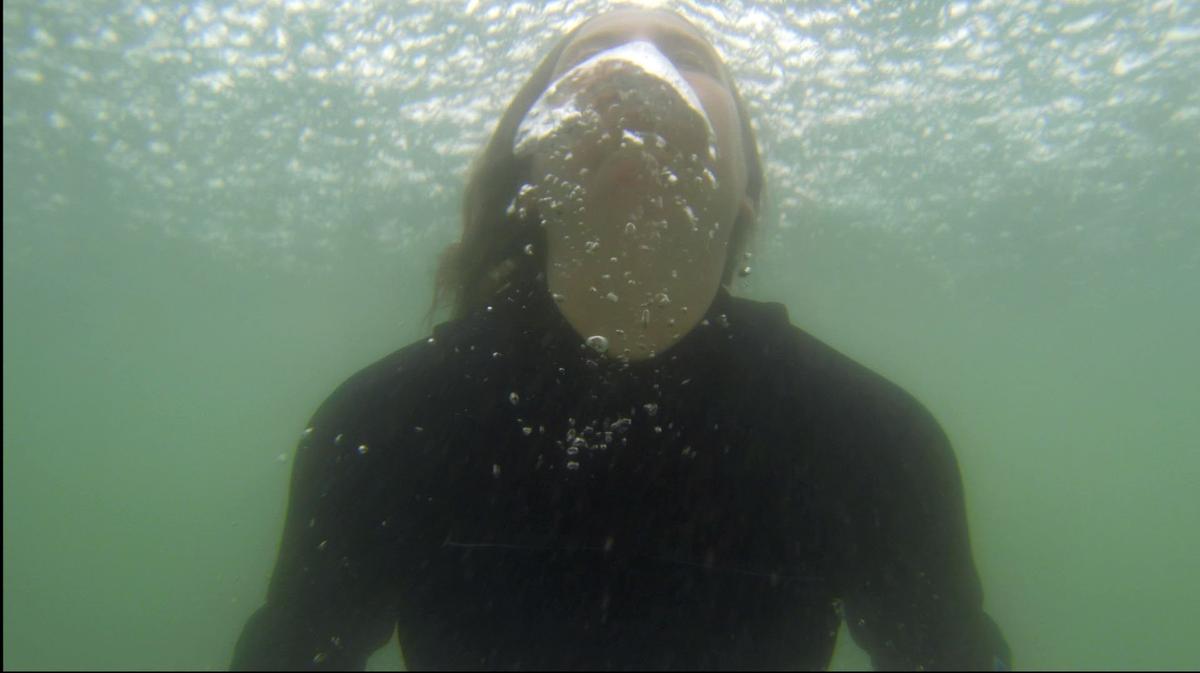 Alex is underwater wearing a wetsuit in a green ocean. She is coming up for air and is exhaling creating large bubbles in the water