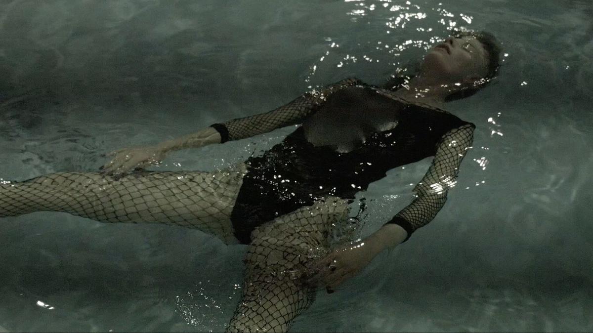 A person wearing a black swimsuit and full body fishnet is floating on their back in a swimming pool. Their head is back, floating, and eyes are closed.  they have black nail polish on. Their body is submerged apart from their chest which is slightly above water.