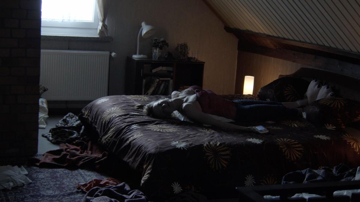 In a messy, dimly-lit bedroom, a women lies on the bed with her back arched and feet on the pillows.