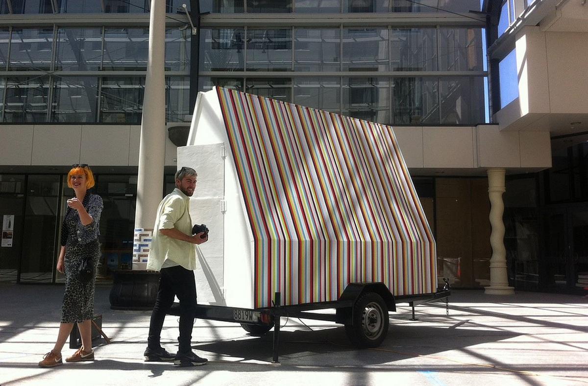 Two people stand next to a large house shaped object which is sitting on a trailer. The house or object is large enough that a person could potentially go inside, it is painted thin precise stripes or black, red, green, pink, blue and baby blue. The two people standing next to the object look happy and excited in the sun coming in from the windows.