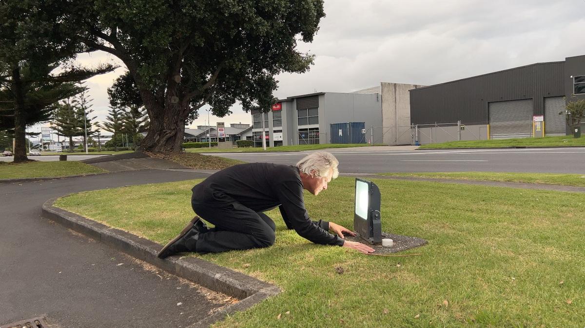 A man kneels on a grass verge and peers into a lamp.