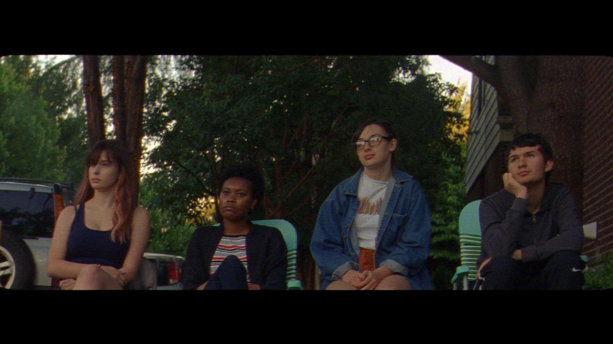 Four teenagers sit outside on the footpath in deck chairs staring to the left of the camera. Behind then are parked cars, the side of a brick building and green trees. They look dreamy, maybe a bit bored but content.