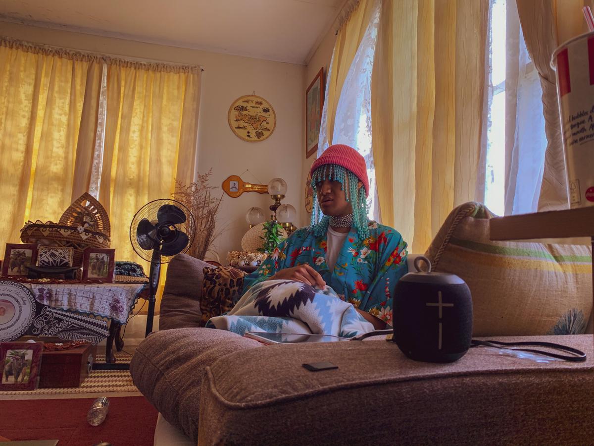 A person sits in a lounge room on a comfortable couch wearing a pink beanie with jewels hanging out of from the beanie. They are listening to something on their Bluetooth speaker