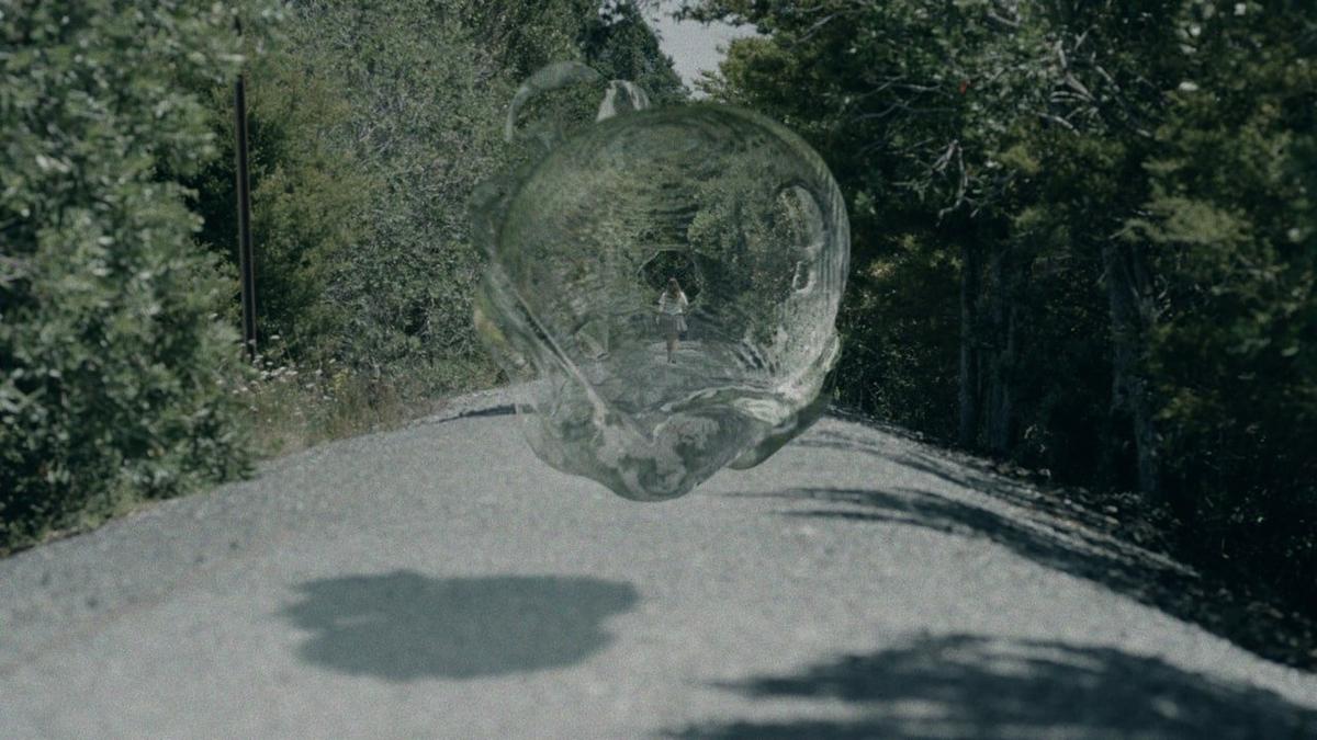 A large water bubble looms over a rural bush road, in the distance a female figure walks with her back to us