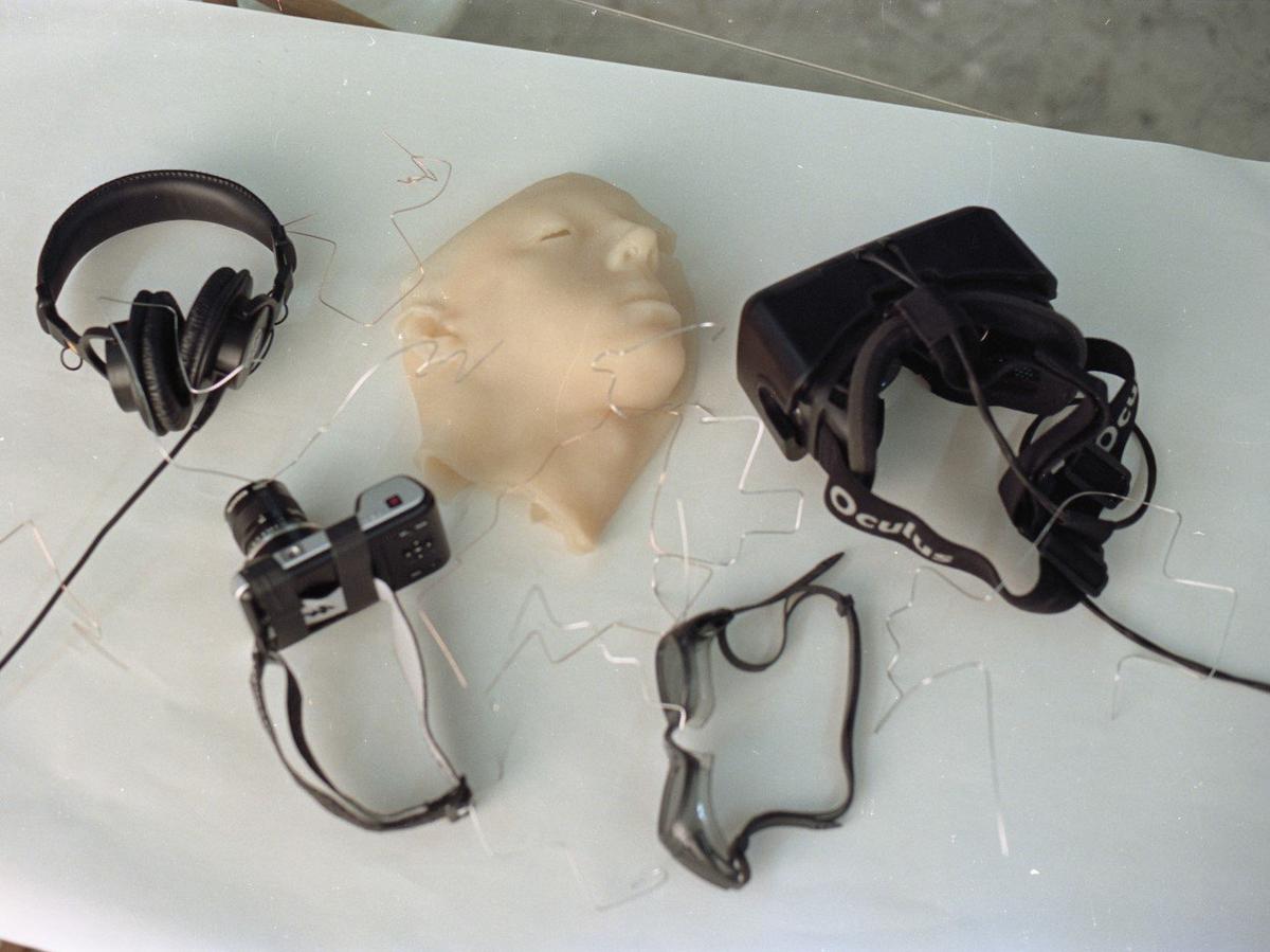 Headphones, oculus, camera, goggles and a plastic face are laid out on a clean white table