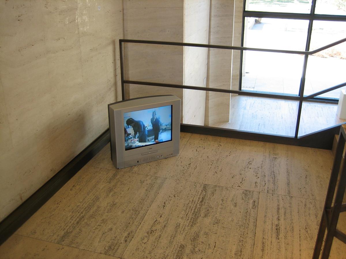 A small video monitor sits on the floor in the corner of what appears to be a stairwell. The monitor screen is in the early television ratio of 4:3, as opposed to the contemporary landscape format of 16:9. The monitor is an installation of Julian Dashper's video work Studio Songs (1998), installed at Sheldon Museum, Lincoln, Nebraska (2006).