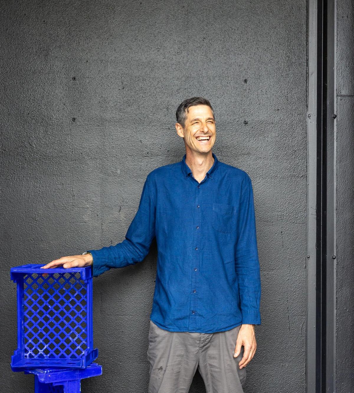 A grinning man in a blue shirt stands against a grey wall; his hand is resting on a stack of blue plastic crates.
