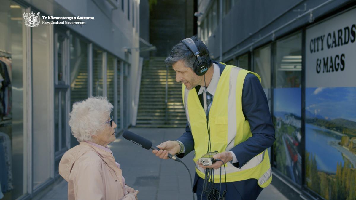 A tall man in a suit and high visibility vest interviews a comparitively dimunutive old lady on the street.