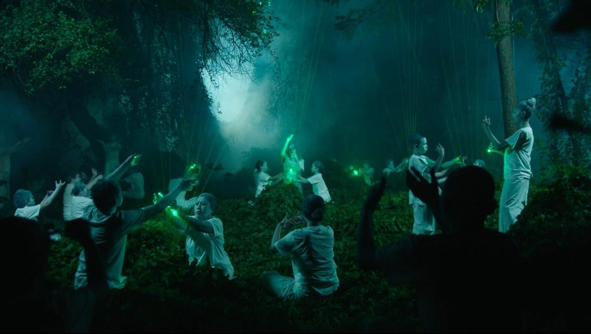 A group of people in white clothing gather in a semi-circle in a forest. Some appear to hold artificial green light. The scene is misty and ritualistic.