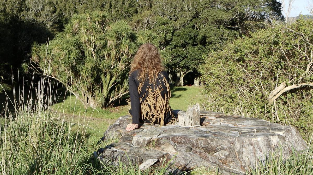 A person sits on a tree stump with their back to camera, they face trees and the sun is shining. They wear adornment made partly from tree leaves.