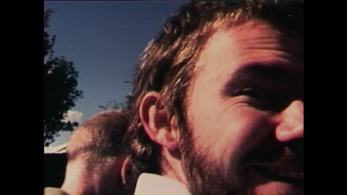 A man's face turns and smiles to the camera; another man's head is visible behind him