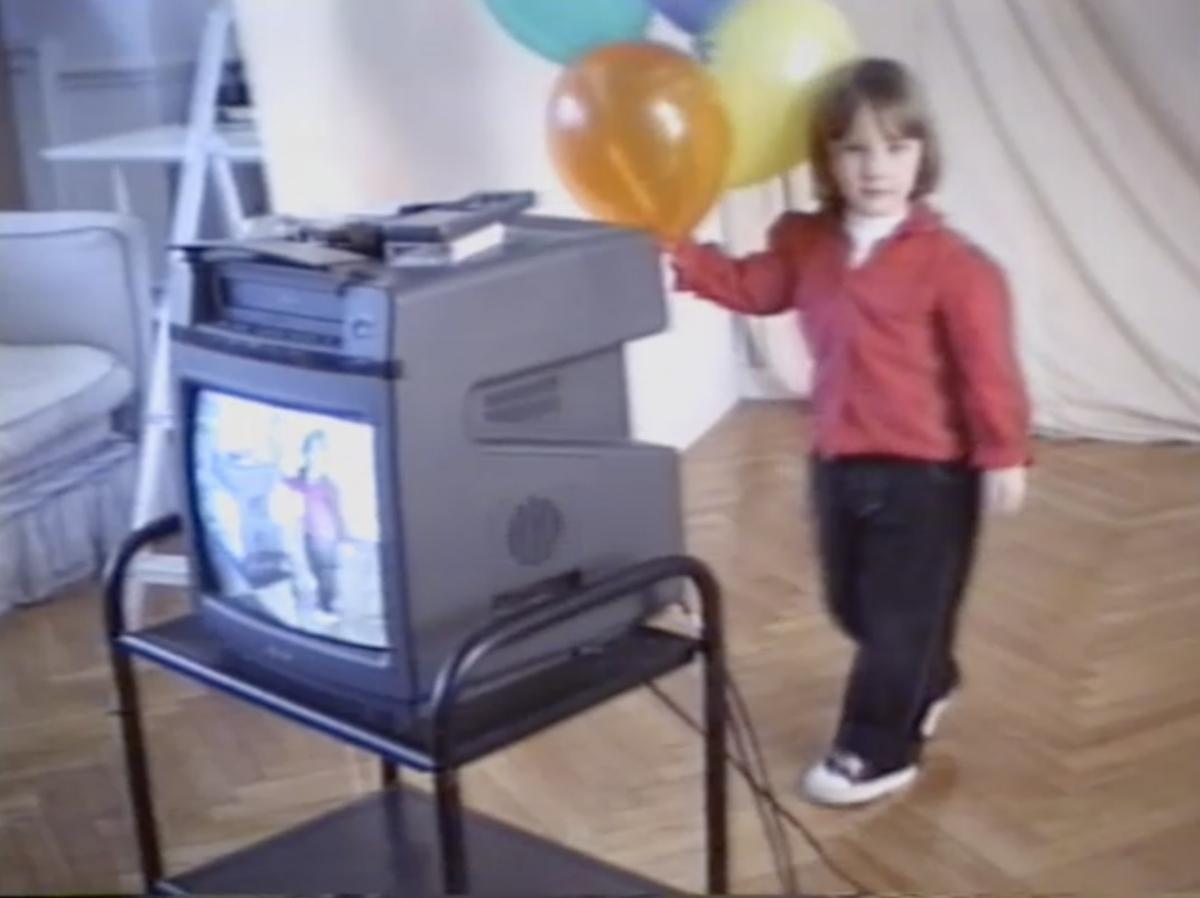 A blurred home video shows a young person holding balloons while a nearby TV monitor shows the same image, indicating she is being videoed live.