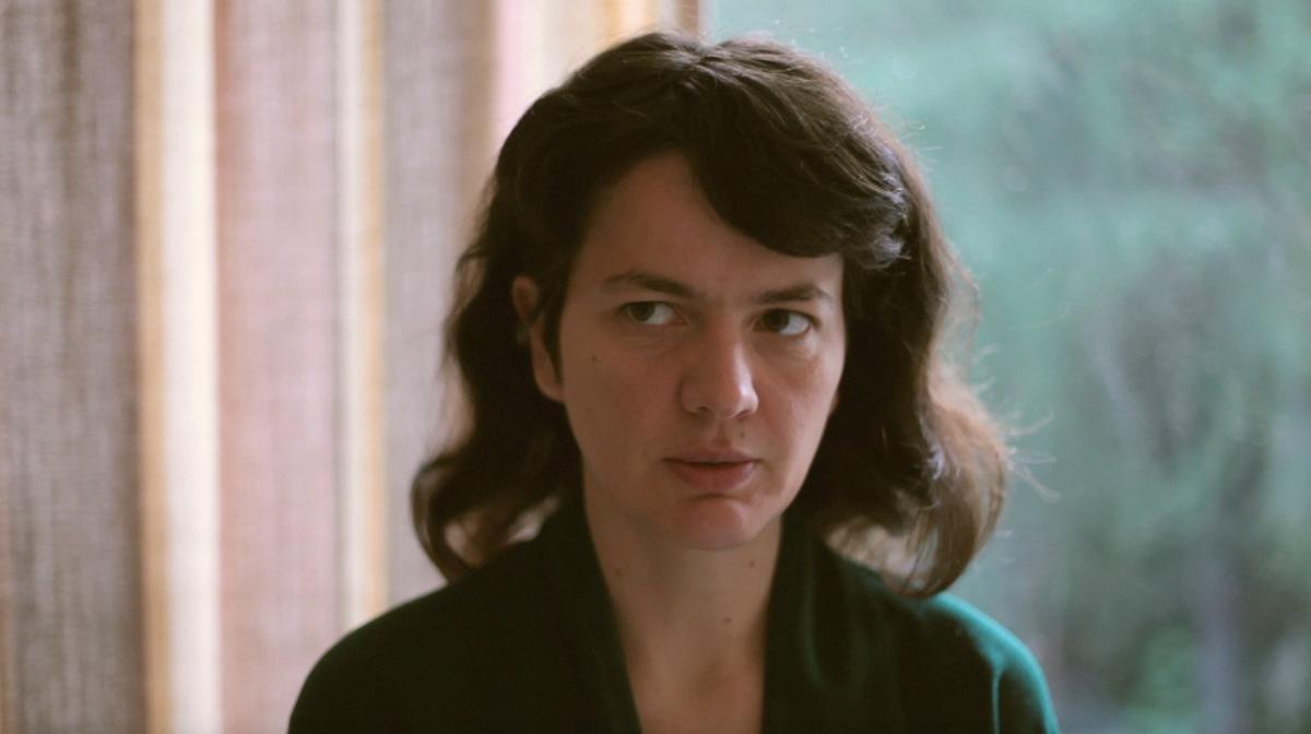 A person with dark hair sits in a domestic setting facing the camera. Their eyes look to something offscreen. The background is blurred, but shows, at left, a pastel coloured curtain. At right, we see a window through whichappears to be nature.