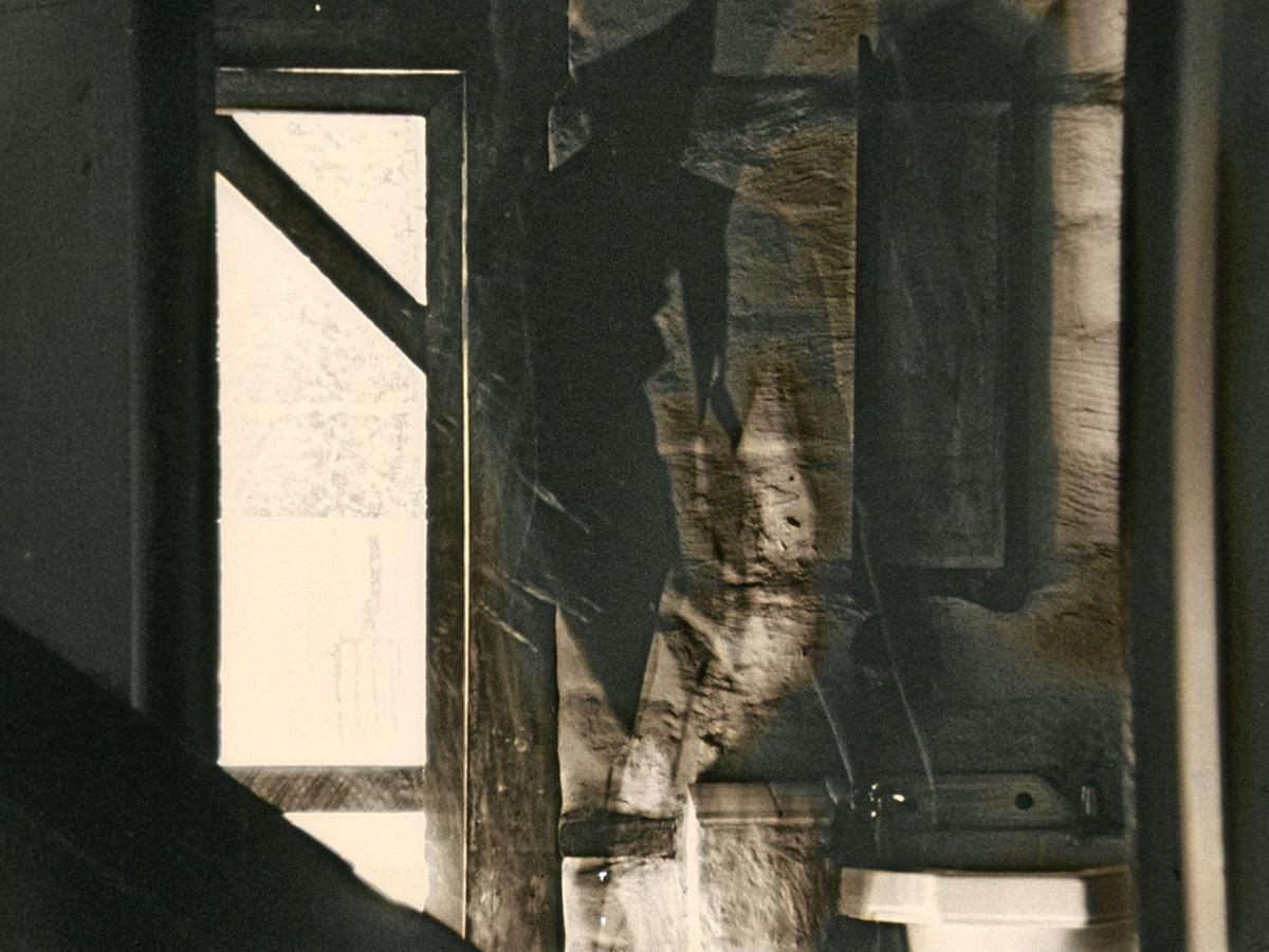 An interior image of an old mud brick hut in St Bathans in Otago. At left is a door.  The centre of the image appears to show the partial image of a human figure hanging in space. At bottom right is a white sink. The still is from a film shot on black and white 16mm film. The film has been rewound and re-exposed multiple times. Each shot is from the same vantage point. The result is a thick layering of interior architectural lines and traces of movement in the room.