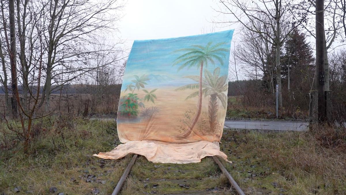 A painted background of palm trees, sand and sea is hung over a railroad track in the middle of what appears to be rural Europe. The sky is grey. The railway track points towards a road, indicating the track is now unused.