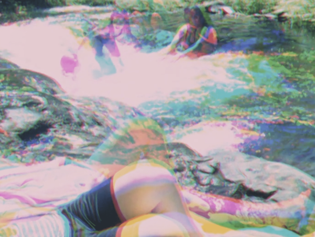 People play near a river or waterfall, laying near the water or swimming. The image has been made with a photographic technique called three colour separation, in which the scene is filmed three times, and each negative given a red, blue and green primary colour. These negatives are layered on top of each other, and combined into one image, which appears to show an ‘after-image’ of different movements recorded in each shot.