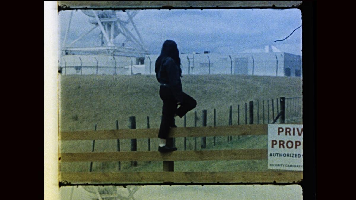 A person stands on a rural fence looking at an astronomy observatory behind a wire security fence