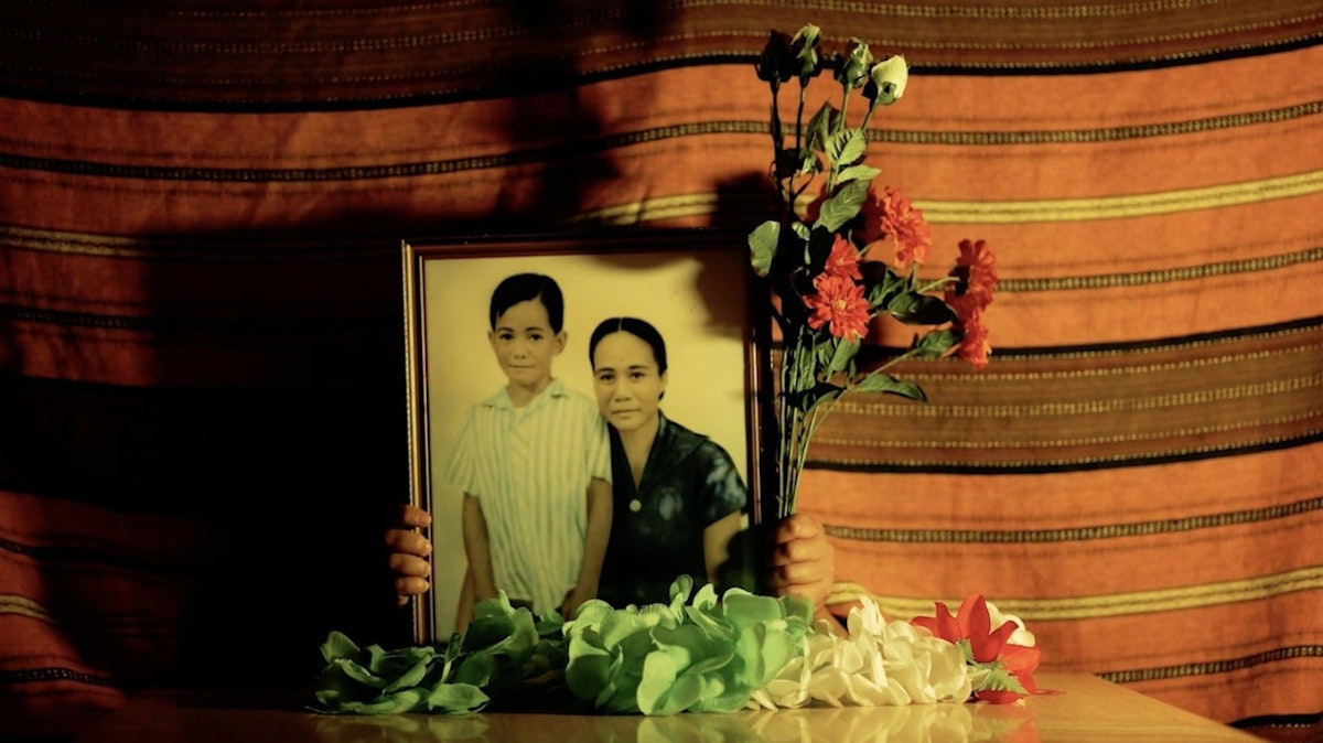A portrait of a woman and a young boy sit on a table surrounded by artificial flowers