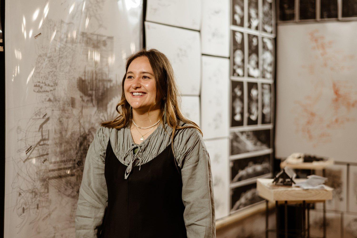 Portrait of a smiling young woman; an exhibition or active studio can be seen in the background.