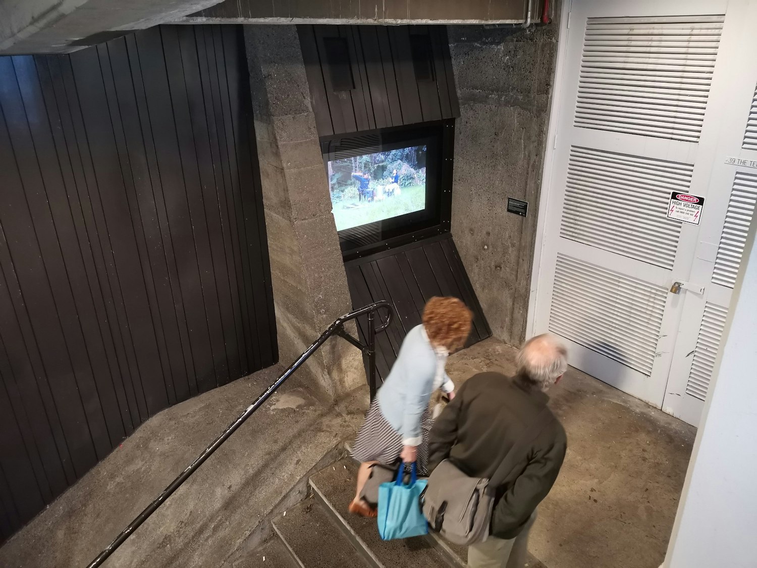Two people are walking up the steps of Mason Lane, a central city laneway between The Terrace and Lambton Quay. They are walking past Masons Screen which is displaying a video work by Arapeta Ashton and Wai Ching Chan.