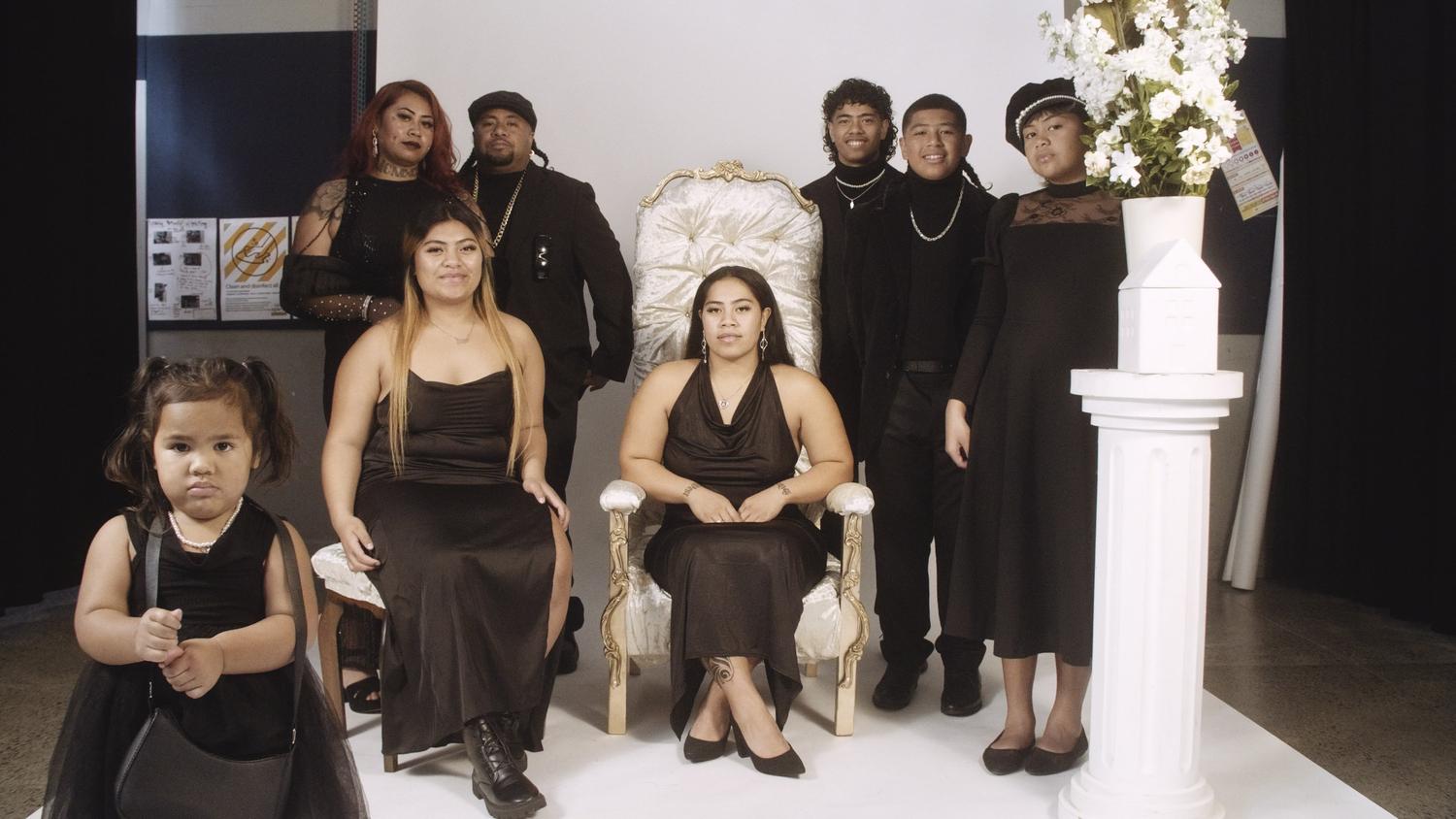 A studio portrait of a Pasifika family dressed in formal attire, facing the camera and sitting or standing on a studio platform. The family spills outside the photographers staged set revealing the artificial construct of the studios